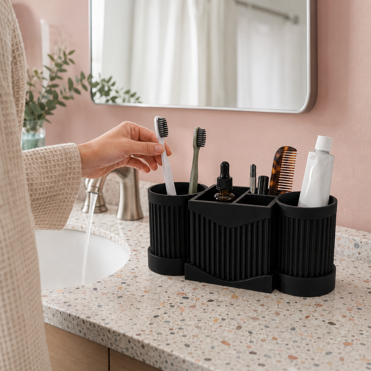 A black silicone bathroom organizer with three cylindrical compartments sits on a speckled countertop beside a sink. A person's hand is placing a toothbrush into the left compartment, with toothbrushes, a comb, a small bottle, and a tube of toothpaste stored inside.