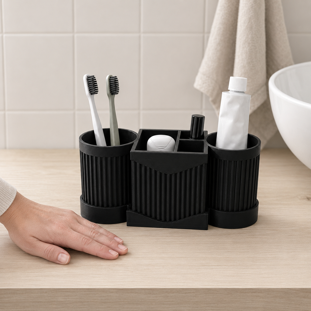 A black 3-in-1 bathroom organizer set on a light wood countertop, holding toothbrushes, a toothpaste tube, and a small white item, with a person's hand resting nearby and a tiled wall and towel in the background.