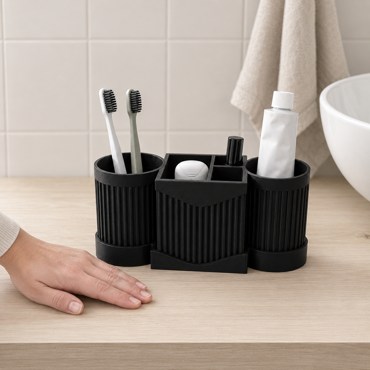 A black 3-in-1 bathroom organizer set on a light wood countertop, holding toothbrushes, a toothpaste tube, and a small white item, with a person's hand resting nearby and a tiled wall and towel in the background.