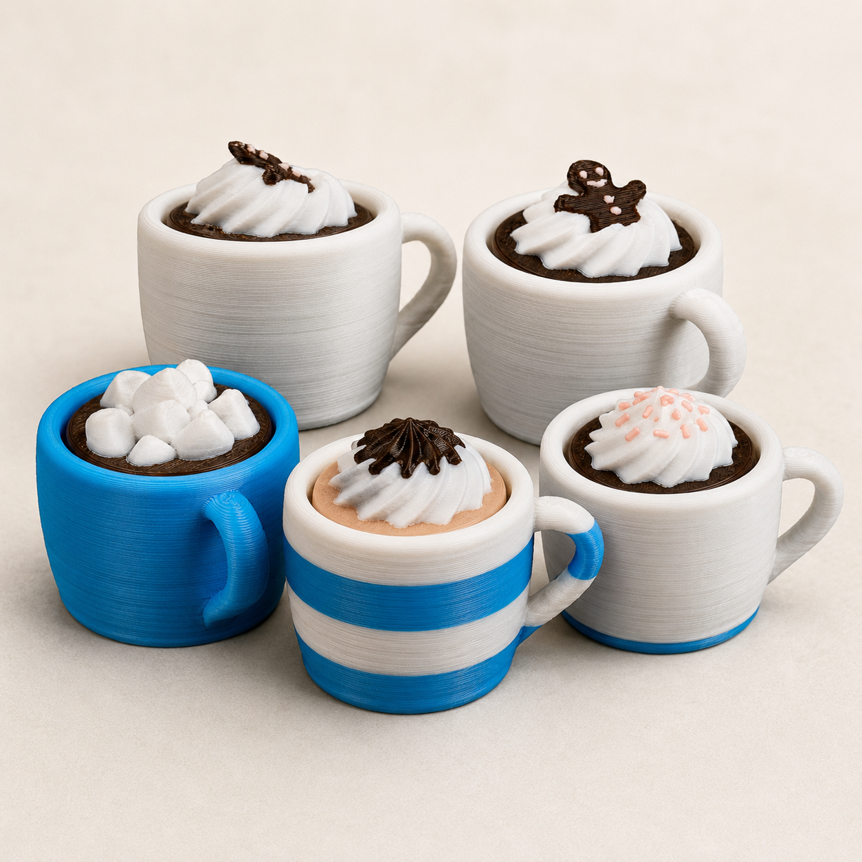 Five miniature hot cocoa cups in various colors and designs, arranged on a light beige background. The cups include white, blue, and blue-and-white striped bodies, filled with dark chocolate and topped with white whipped cream, chocolate drizzle, marshmallows, and pink sprinkles.