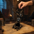 A fantasy tabletop scene in a dimly lit stone room. A sculpted tree-like object with skull faces sits on an old map, with a human hand placing a die near the top.