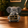 A gray elephant figurine holding a sign that reads 'TUSK ME IF I CARE' sits on a wooden table in a warm, dimly lit room with a dark green wall and blurred background elements.