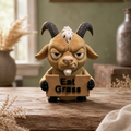 A cartoon goat figurine with black horns and a white beard, holding a cardboard sign that says 'Eat Grass', displayed on a rustic wooden surface with dried floral props and a soft-focus background.