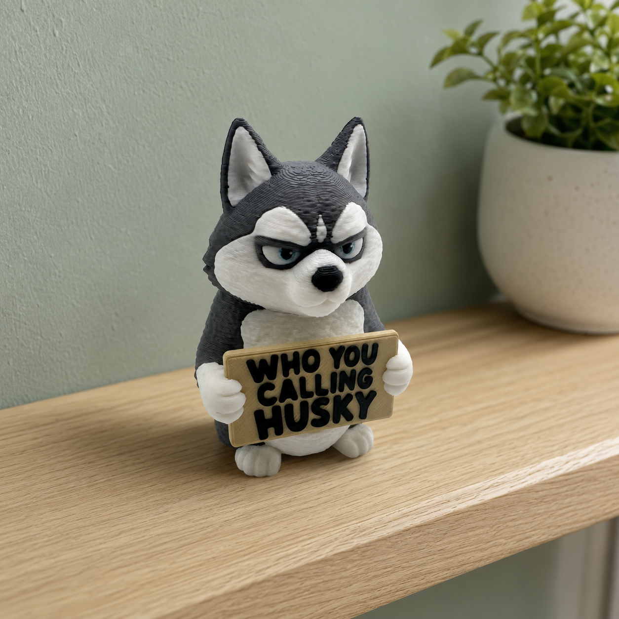 A small husky figurine sitting on a light wood shelf, holding a sign that reads 'WHO YOU CALLING HUSKY'. The background shows a soft-focus green wall and a potted plant.